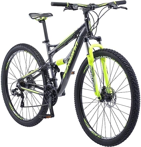A blue and yellow Schwinn Traxion mountain bike with front and rear suspension, featuring large 29-inch wheels, disc brakes, and a front fork lockout mechanism for versatile riding on various terrains. The bike is positioned outdoors on a dirt trail, surrounded by green foliage.